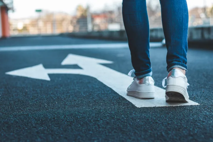 A shot of a person's feet on a road walking toward fork in the road icon painted on the street.